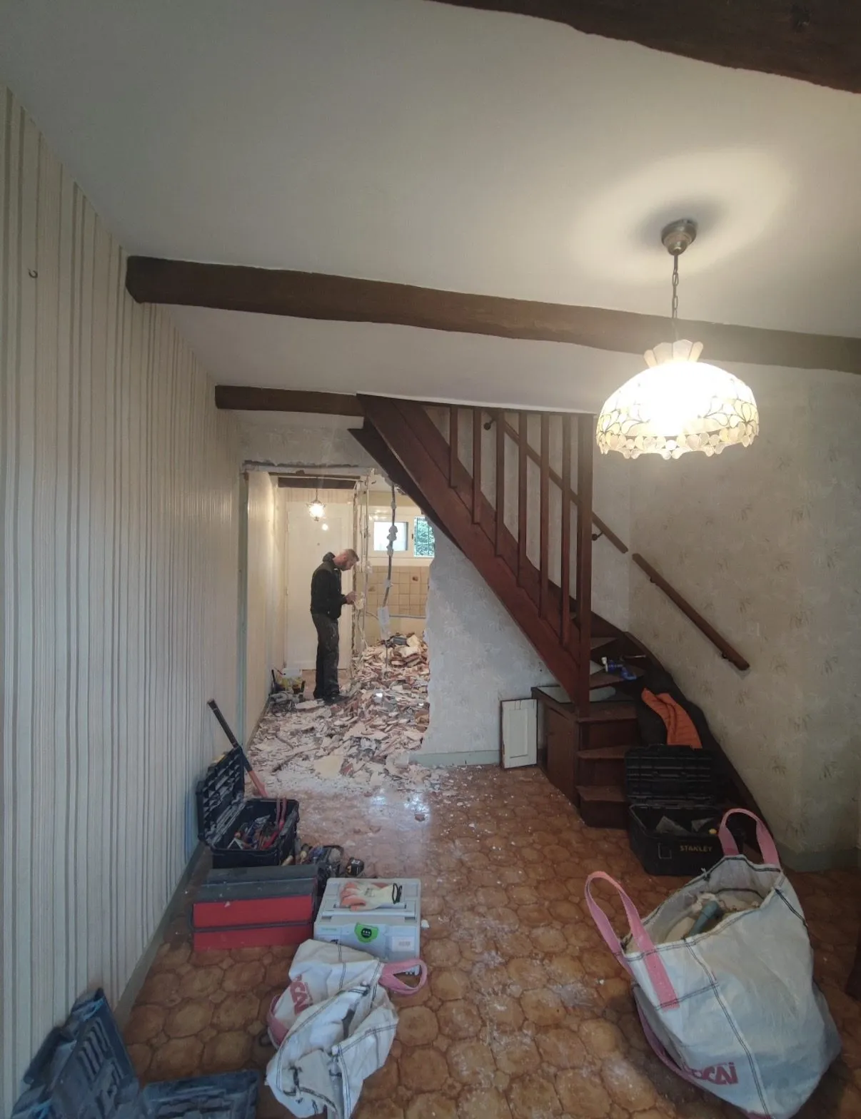 Démolition d'une cloison dans le couloir d'une maison ancienne, sous un escalier en bois, avec gravats au sol