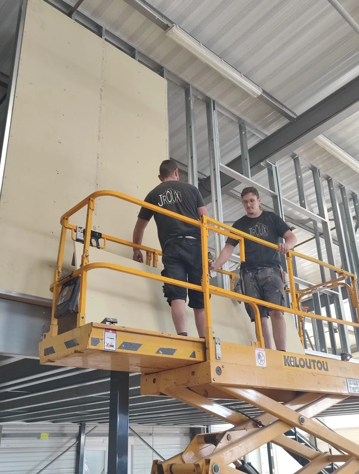 Deux compagnons de JRoux Rénovation posant une plaque de plâtre sur nacelle dans un local professionnel