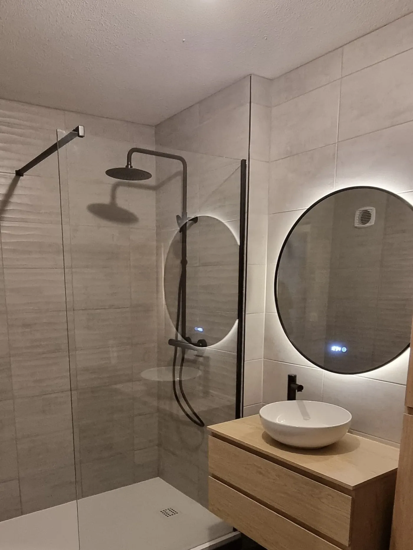 Salle de bains avec douche à l'italienne, paroi fixe, robinetterie noire, miroir rond rétroéclairé et vasque bol sur meuble chêne